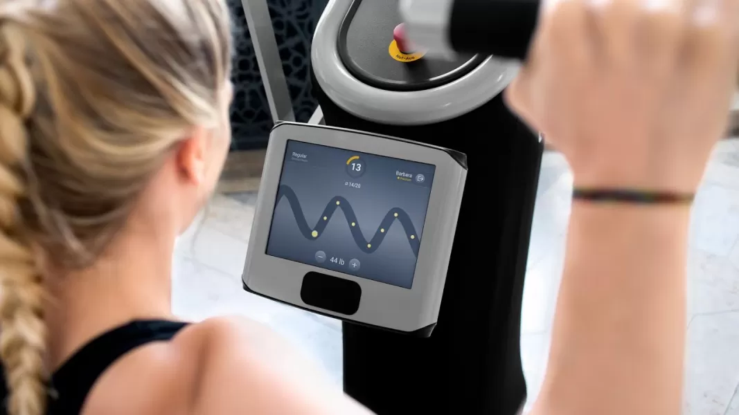 A woman looks at an eGym screen that displays a ball moving through a pathway, measuring her strength on the shoulder press