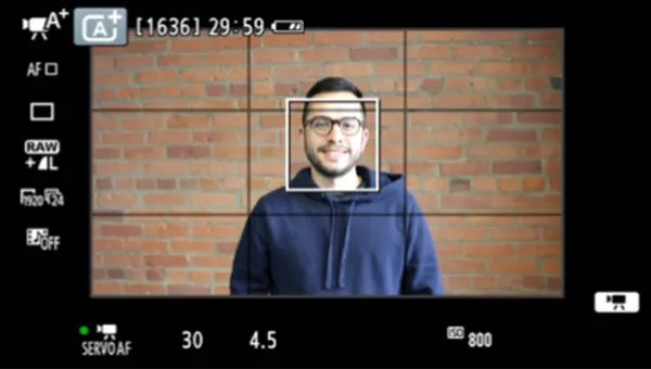 A man stands smiling with a brick wall behind him. The camera display icons see in a square formation all around him