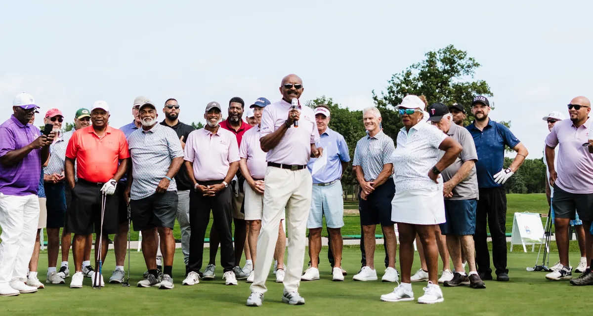 A group of golfers and spectators stands on a green course, with several holding clubs and a caddie visible at the right edge.