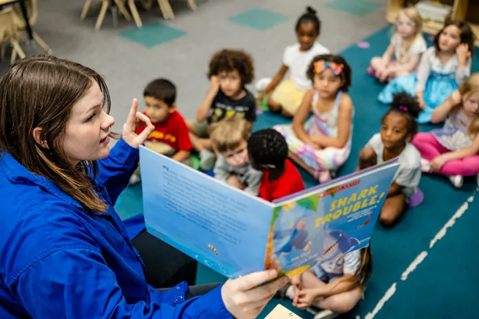 A teacher reads a storybook to a group of children sitting on the floor in a classroom. The book cover shows a shark, and the children are engaged and listening attentively.