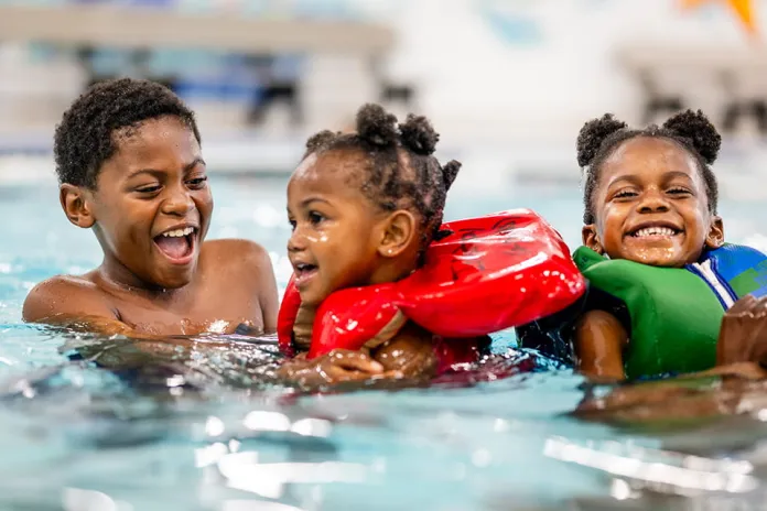 Three children enjoying swimming in a pool. Two girls wear flotation devices, smiling and playing with a boy nearby, all surrounded by clear water and a colorful, child-friendly environment.