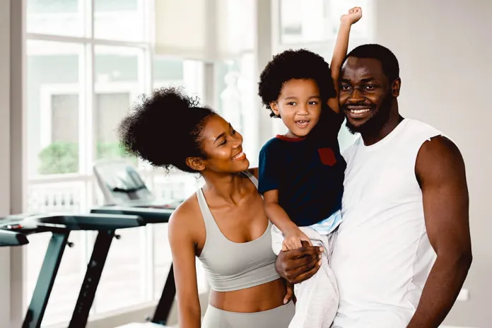 A woman in activewear and a man holding their smiling child in a gym setting with exercise equipment in the background and large windows providing natural light.