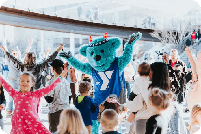 A person in a blue furry mascot costume and YMCA shirt stands with raised arms among children and adults outdoors under a bridge.