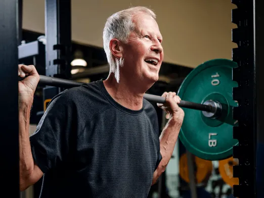 An older man in a dark T-shirt holds a barbell with green weight plates across his shoulders in a gym.