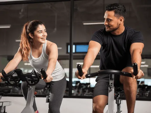A woman in a white tank top and gray leggings and a man in a black T-shirt and shorts ride stationary bikes in a gym with large windows behind them.