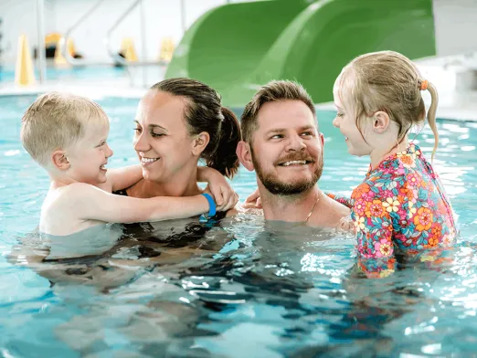 A smiling family of four stands in an indoor swimming pool, with two young children holding onto each parent and a green waterslide in the background.