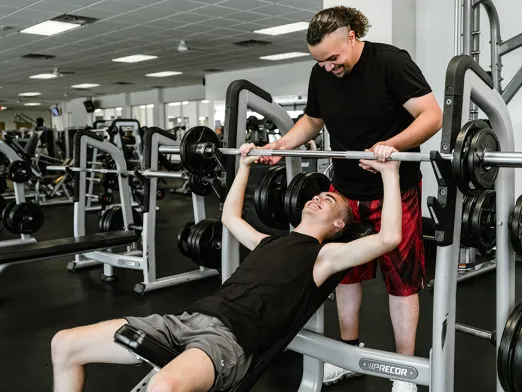 A man spots another man doing a bench press on an incline bench, with weight machines and mirrors in a gym background.