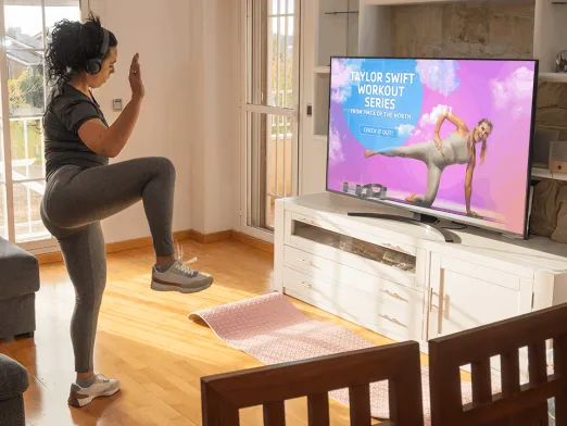 A woman in workout clothes balances on one leg in a living room while following a Taylor Swift workout series on a TV, with a yoga mat on the floor beside a white media console.