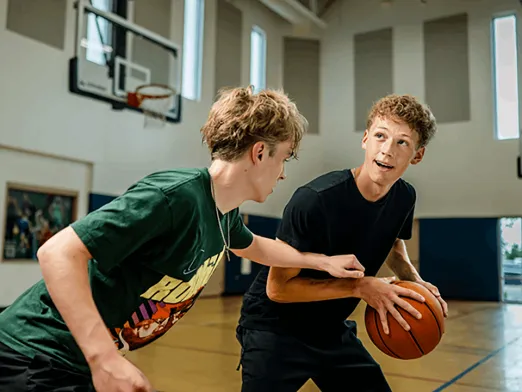 Two young men play basketball in a gym, with one dribbling a ball while the other reaches in to defend near the hoop.