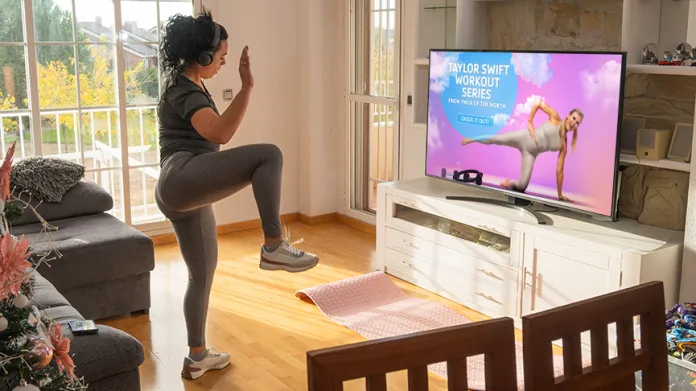 A woman wearing headphones is exercising in a brightly lit living room. She is performing a workout move with one leg raised. A television displays the 'Taylor Swift Workout Series' with a fitness instructor demonstrating an exercise.