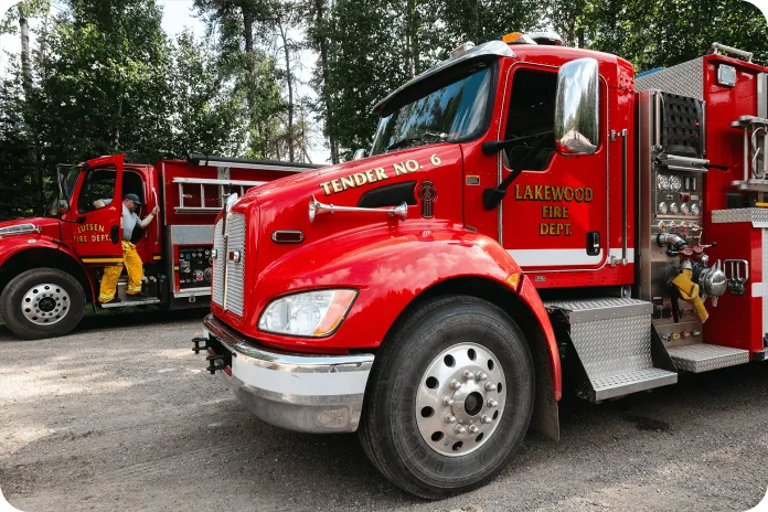 A red fire truck labeled 'Tender No. 6' is parked on a gravel road. To the left is another fire truck from the 'Lakewood Fire Dept.' with a firefighter in a yellow uniform standing beside it, surrounded by green trees in the background.