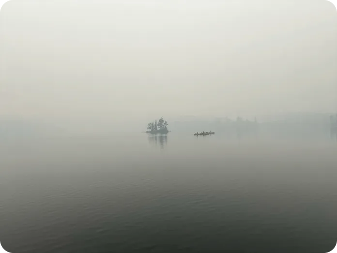 A smokey lake scene featuring a small island with trees in the center. A canoe with two people is visible on the water, surrounded by a misty atmosphere and a serene reflection on the calm surface of the lake.