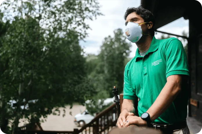 A man wearing a green polo shirt and a face mask stands on a wooden balcony, leaning on the railing. In the background, there are trees and parked cars visible in a natural outdoor setting.