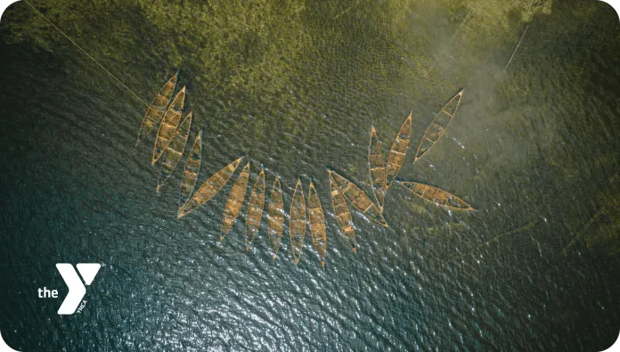 An aerial view of several wooden boats arranged in a formation on the water, surrounded by a greenish shoreline. The boats vary in size and orientation, and the sunlight reflects off the water's surface, creating a shimmering effect.