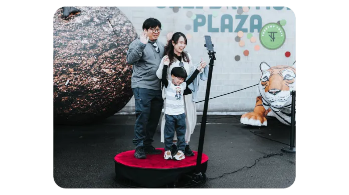 A family of three stands on a red circular platform taking a selfie with a smartphone on a stand, with a large decorative sphere, tiger figure, and “PLAZA” sign in the background.
