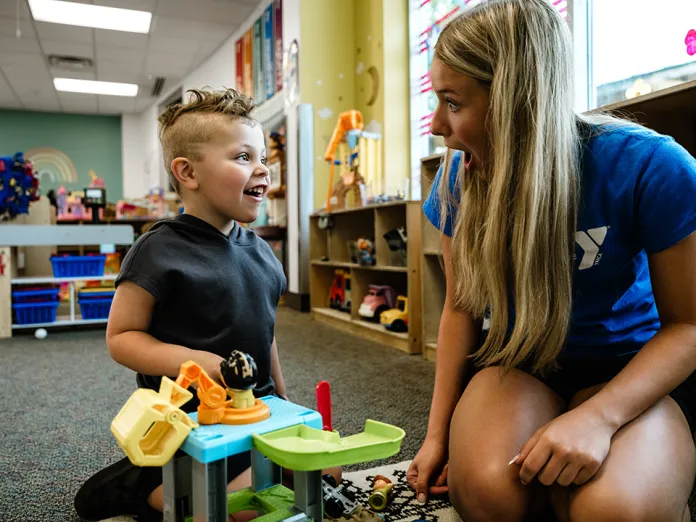 A young boy and a woman kneel facing each other on a classroom carpet, with toy blocks and play equipment beside them.