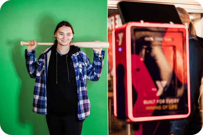 A woman in a blue plaid shirt holds a wooden bat across her shoulders in front of a green screen, with a red-lit photo booth or arcade cabinet beside her.