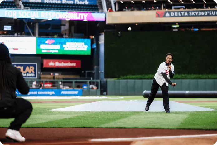 D'Mario in a Twins jersey throws a baseball on a stadium field, to his mentor who is out-of-focus in the foreground with sponsor signs in the stands.
