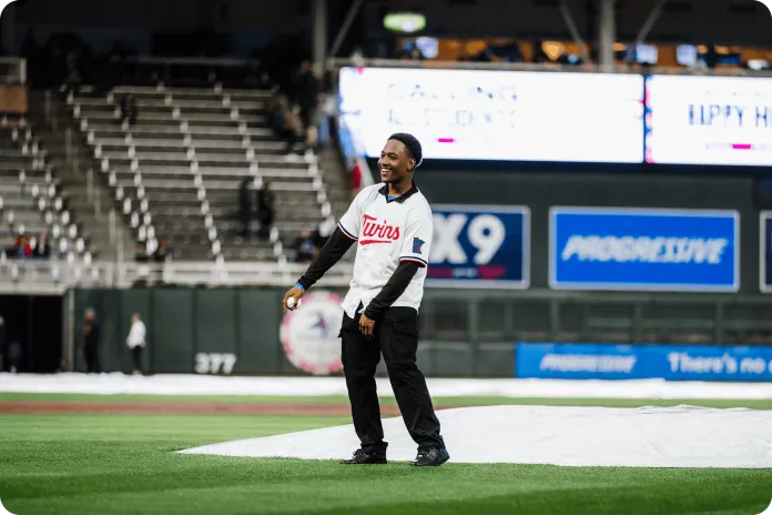 D'Mario in a Minnesota Twins jersey stands on a baseball field near a white tarp, with stadium seats and video boards in the background.