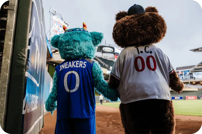 Mascots from YMCA of the North, Sneakers, and T.C Bear, holding hands on Target Field at a Twins baseball game.