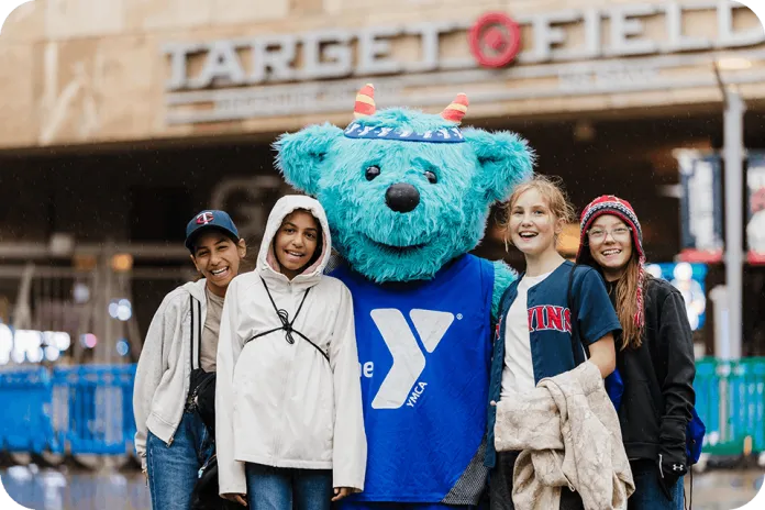 Four children stand with the YMCA of the North's blue furry mascot, Sneakers, in front of a Target store entrance, with blue barriers and a wet pavement in the foreground.