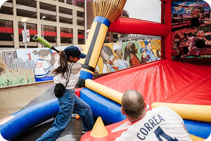 A child in a cap plays on an inflatable obstacle course with red and blue walls, while an adult in a  jersey watches nearby, with murals and a building facade in the background.
