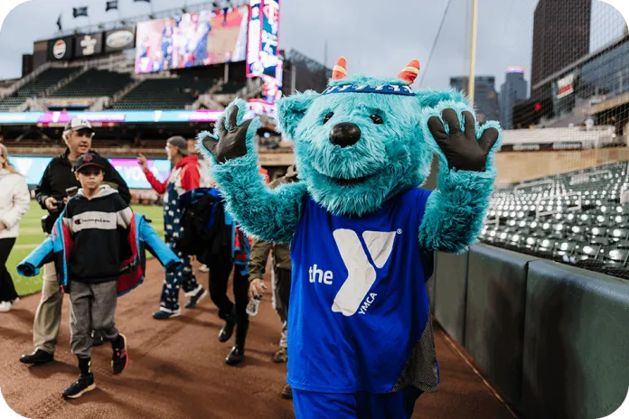 YMCA of the North mascot, Sneakers waving on a baseball field during the children's parade with spectators and stadium seats in the background.