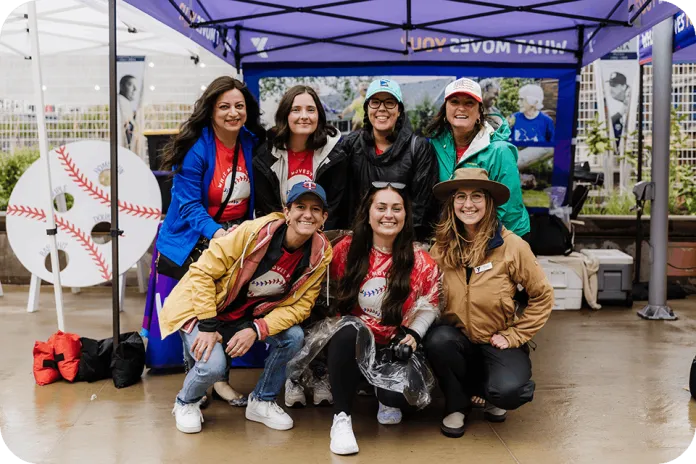 Seven women in baseball-themed shirts pose under a purple canopy at an outdoor event, with a large circular baseball sign and vendor booths in the background.