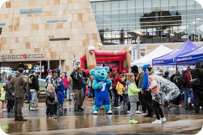 YMCA of the North mascot, Sneakers, stands in a crowd outside Target Field, with vendor tents and a red inflatable entrance in the background.