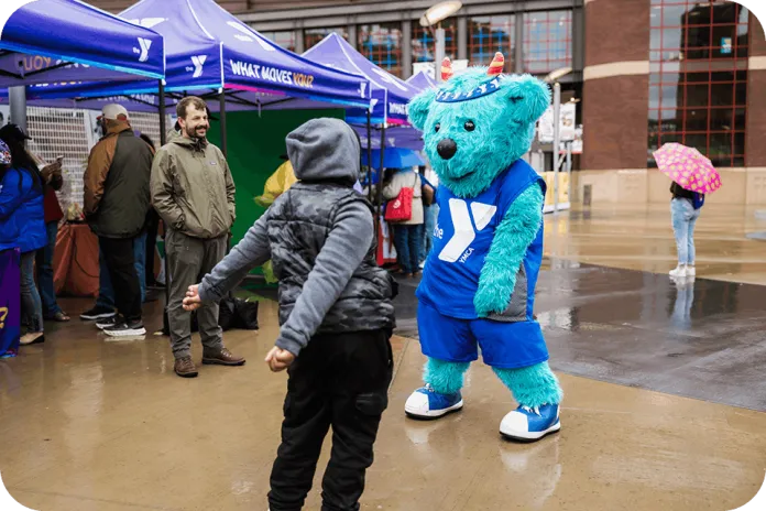 YMCA of the North mascot, Sneakers, wearing a YMCA jersey stands on a wet sidewalk while a child in a gray hoodie faces it, with blue pop-up tents and other people in the background.