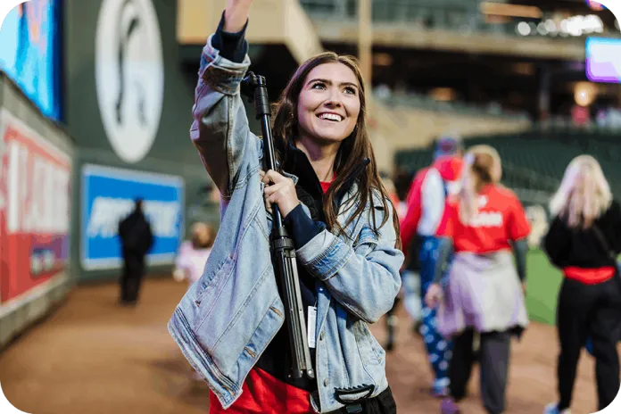 A smiling woman in a denim jacket holds a camera tripod with one raised arm in a stadium concourse, with several people walking in the background.