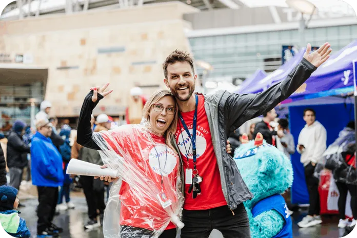 A smiling man and woman in red shirts stand together at an outdoor event, wearing clear plastic rain ponchos and waving at the camera.